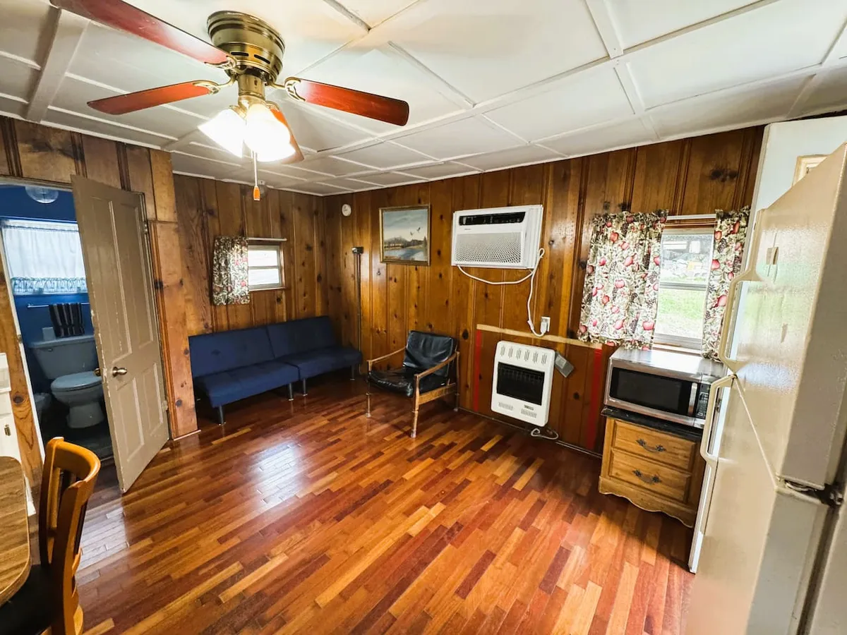 This interior image features a cozy living space with wooden walls, a comfortable sofa, a dining area, and visible kitchen appliances. Natural light enters the space, creating a warm and welcoming atmosphere, ideal for relaxing or socializing after a day at the lake.
