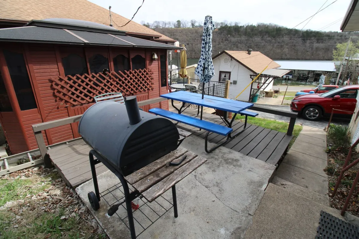 A social and inviting outdoor area with a grill, picnic tables, an umbrella for shade, and several houses in the background. The space is set up for barbecuing, dining, and enjoying time with friends or family outdoors.