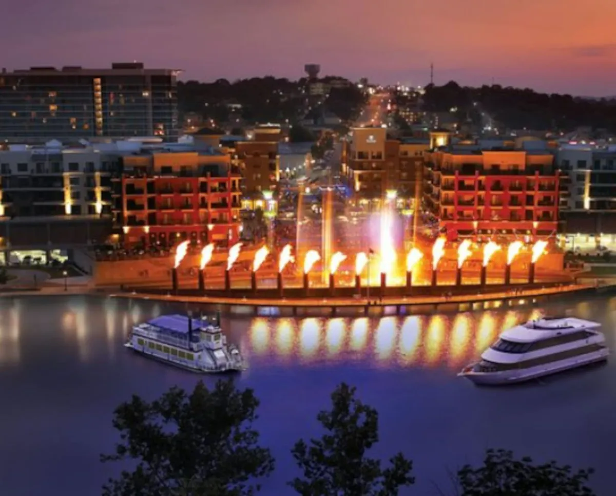 The scene captures a vibrant waterfront area in Branson, featuring illuminated fountains, colorful buildings, and people dining by the water as boats pass by. The festive mood and lively setting suggest a variety of dining options nearby, including the opportunity to enjoy local restaurants.