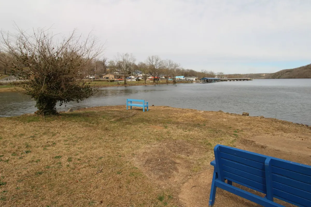 A peaceful waterfront scene with blue benches, tree-shaded spots, and distant houses, inviting guests to relax, picnic, or enjoy food by the water. The tranquil setting highlights another way to enjoy flexible dining—packing a picnic for a scenic lunch outdoors.