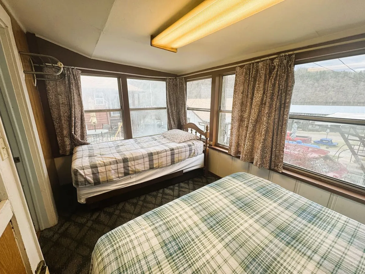 A cozy bedroom featuring two beds with inviting plaid bedding, complemented by large curtained windows that allow plenty of natural light. Wooden furniture adds warmth and a sense of rustic comfort, making the room feel both nostalgic and welcoming.