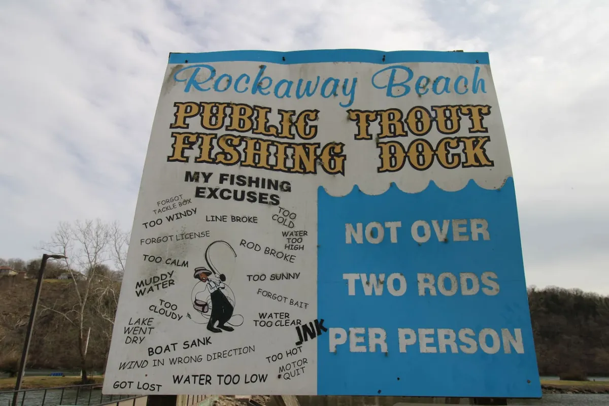 A playful sign marks the 'Public Fishing Trout Dock,' listing humorous fishing excuses and inviting guests to relax, fish, and socialize by the water. The dock and water are visible in the background, underscoring the community aspect of fishing on Lake Taneycomo.