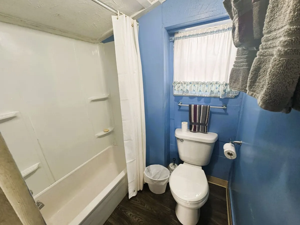 A fresh and practical bathroom with calming blue walls, a spacious shower area, a clean toilet, and neatly arranged towels by a curtained window. The space is designed for both relaxation and convenience, providing all the essentials for guests to refresh after a long day.