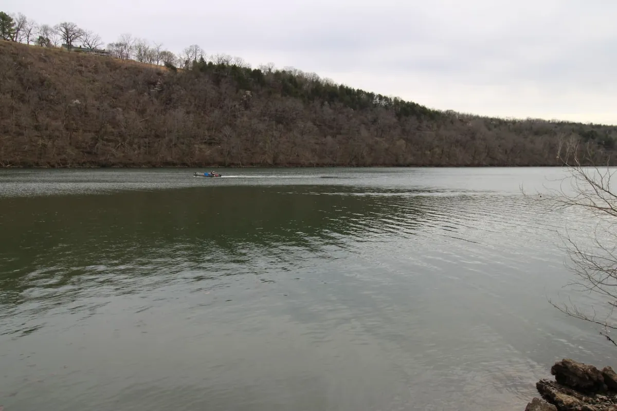 A serene view over calm waters with a kayak or canoe gently floating, distant tree-lined hills reflected in the glassy surface. The shoreline is quiet and natural, perfect for moments of peaceful reflection, quiet boating, or birdwatching.