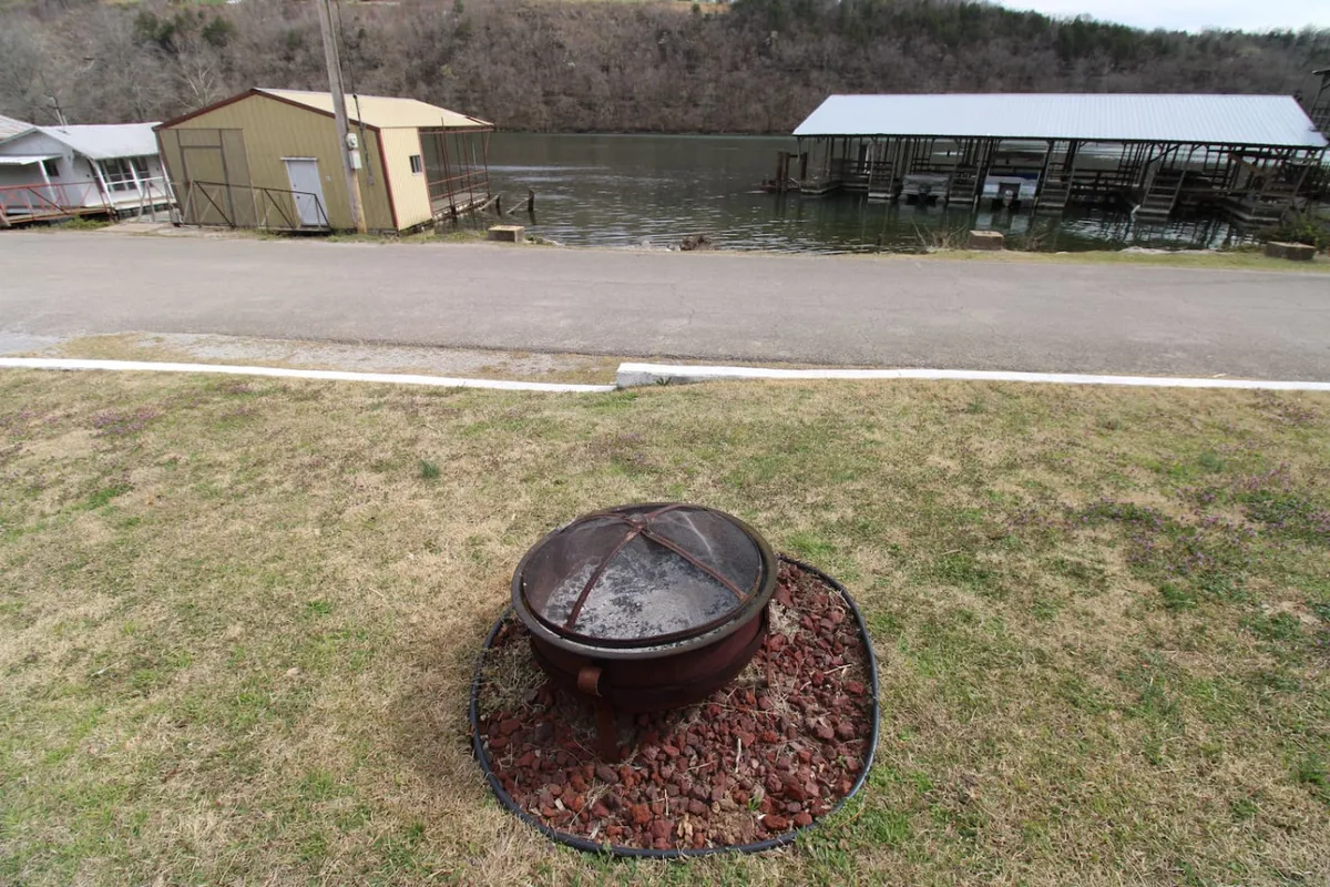 A cozy fire pit area set beside the lake, surrounded by green grass and distant cabins, with the water reflecting the sky. This inviting outdoor space suggests evenings spent by the fire, enjoying nature and unwinding.