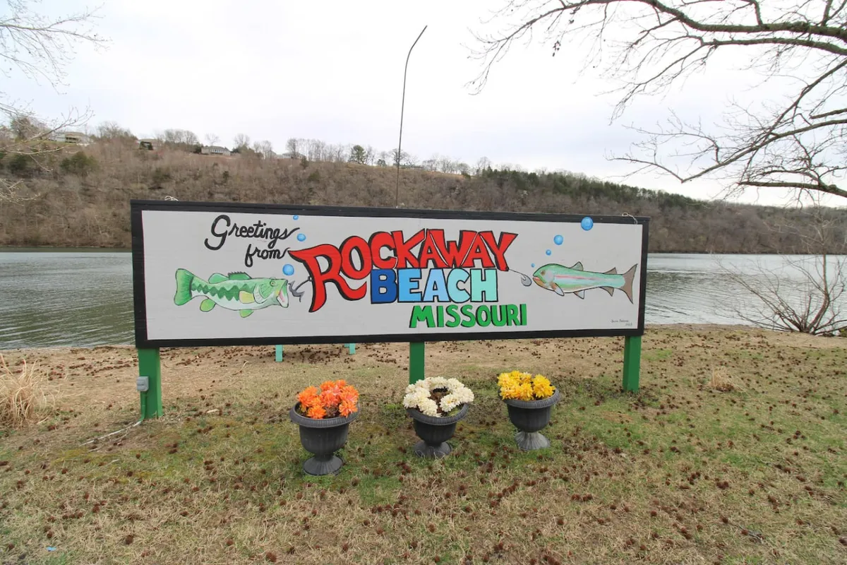 A welcoming greeting sign with cheerful flower pots sits at the edge of a property overlooking the lake, inviting visitors to stop for picnics, fishing, or to simply enjoy the view. The lake glimmers in the background, surrounded by greenery.