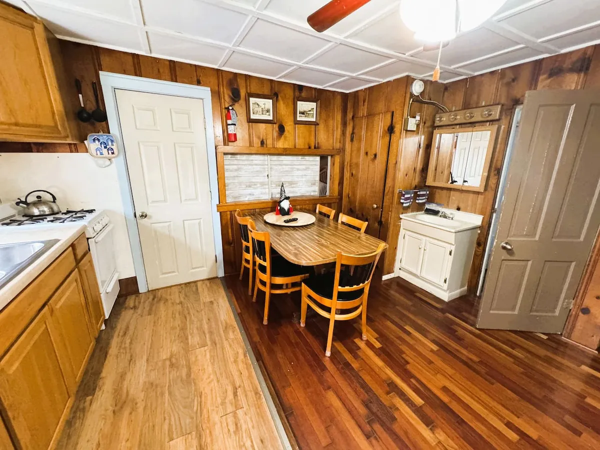 An interior view of a cozy dining area with a wooden dining table, kitchen cabinets, warm wood paneling, and a mirror. The atmosphere is nostalgic and inviting, perfect for sharing meals and connecting with others after cooking in the kitchenette.