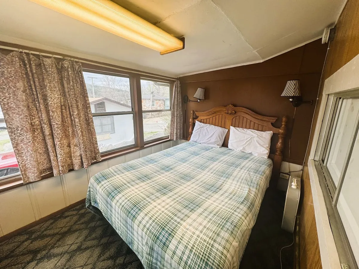 A tranquil bedroom with a soft bed, windows framed by curtains, and warm natural light filtering in. The scene suggests restful relaxation, reading, or quiet contemplation, aligned with the peaceful lakeside theme.