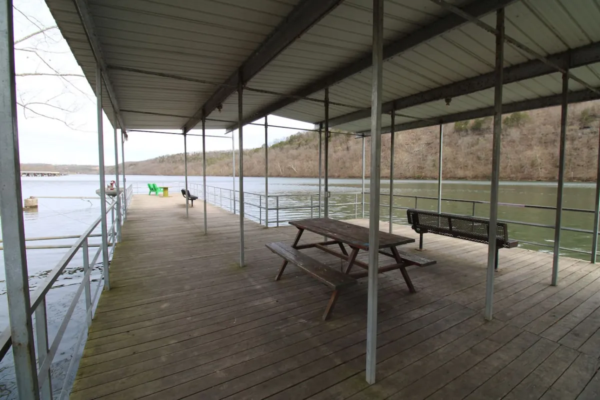 A peaceful lakeside setting featuring a wooden dock extending over calm water with a picnic table, benches, and a green chair. The scene suggests both relaxation and functionality, with clear access to the water for fishing or boating, and ample space for outdoor dining or leisurely gatherings.